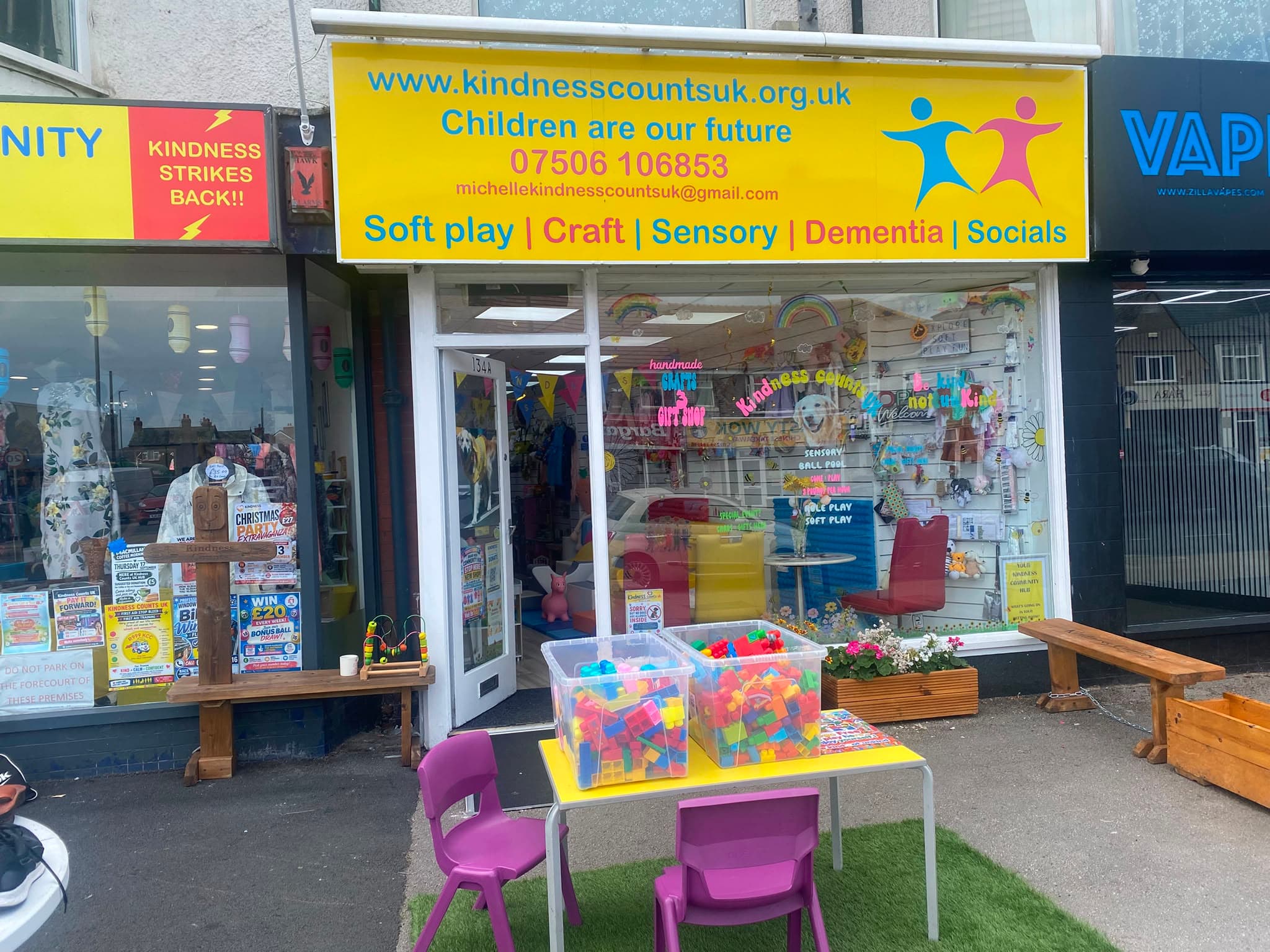 The Kindness Counts UK hub on Victoria Road West, its yellow sign reading 'Children are our future' above a window of soft play and craft, with a children's table and chairs outside.