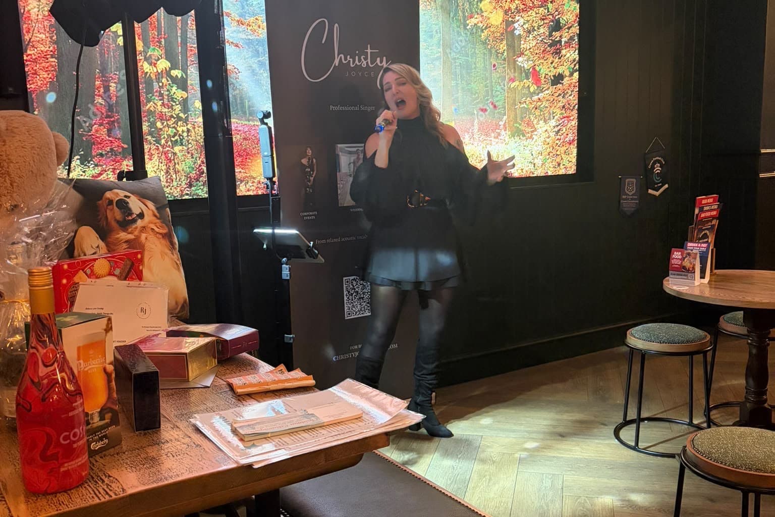 Christy Joyce singing at the Gardeners Arms, beside her banner, with the raffle prizes laid out on a table in front of her.