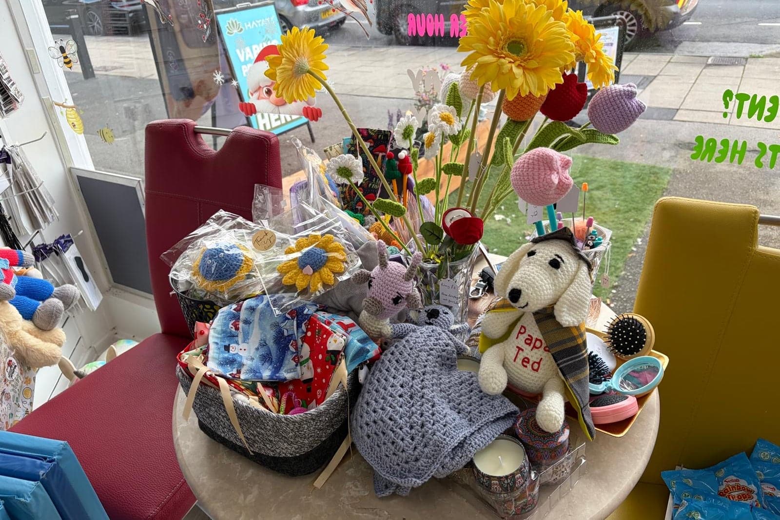 Crocheted flowers, a blanket and Papa Ted on the table by the shop window, with fabric gifts in baskets beside them.