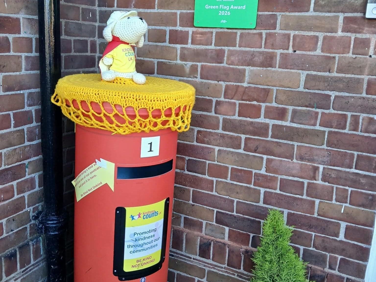 The Kindness Counts UK postbox at Little Thornton Nursery, with a yellow crocheted topper and a crocheted Nic sitting on top.