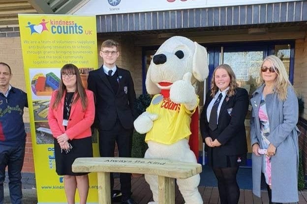 A new buddy bench with “Always Be Kind” carved into it, outside a school — Nic the mascot, two pupils and three of the team standing behind it beside the Kindness Counts UK banner.